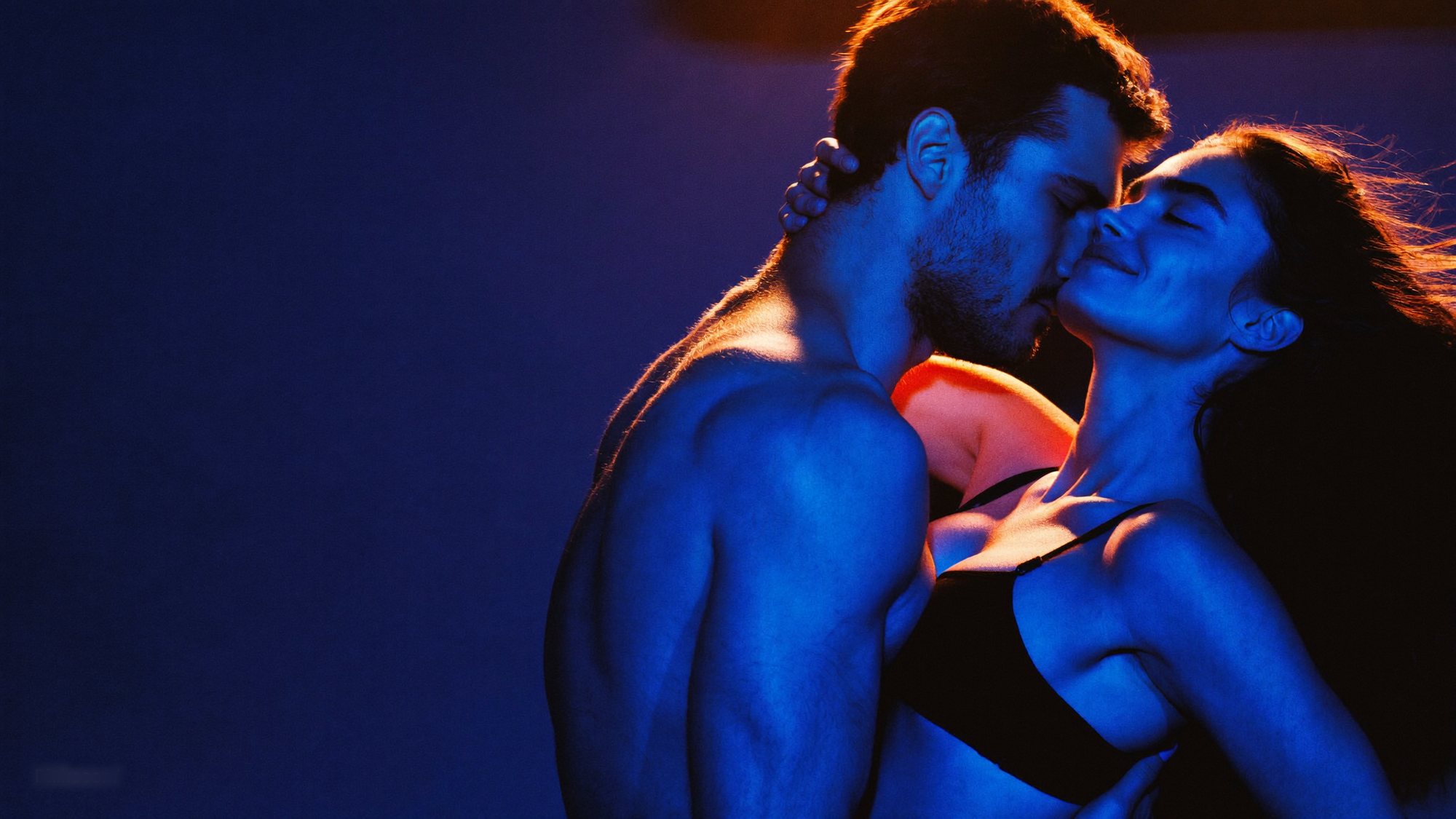 A shirtless man and a woman embrace under blue light