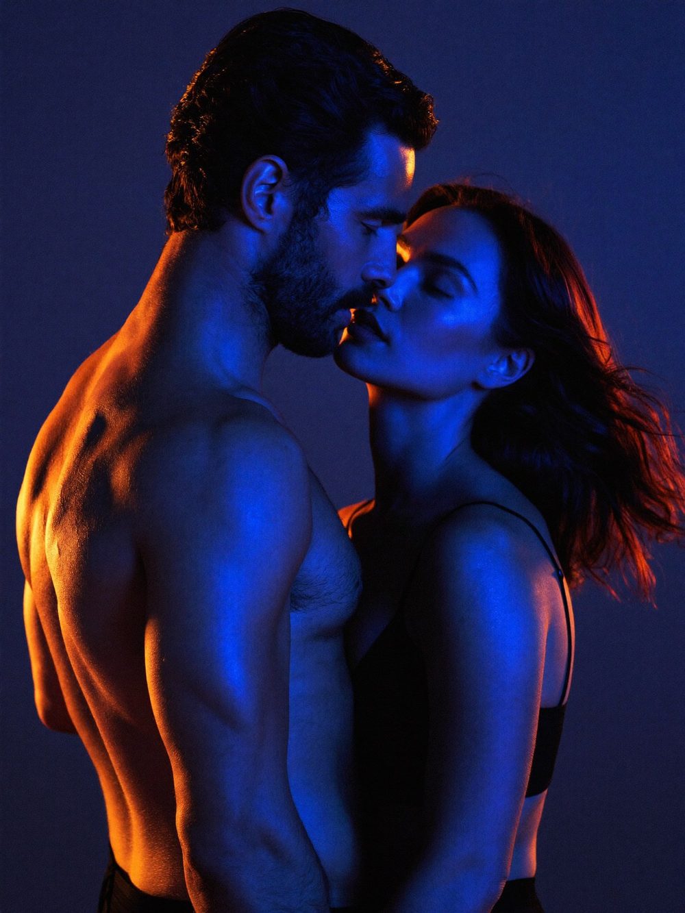 A couple holding each other close with their eyes closed, lit in deep blue with warm amber rim light
