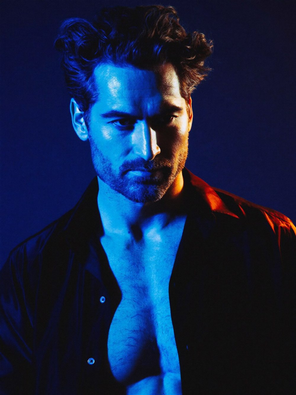 A confident man in an open black shirt under blue light